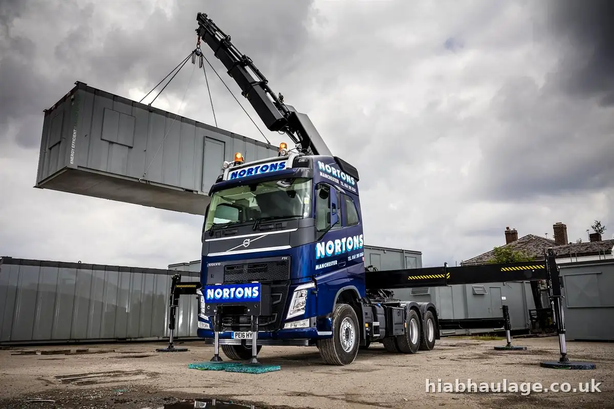 Artic HIAB with its stabilisers out lifting a grey site cabin in the yard