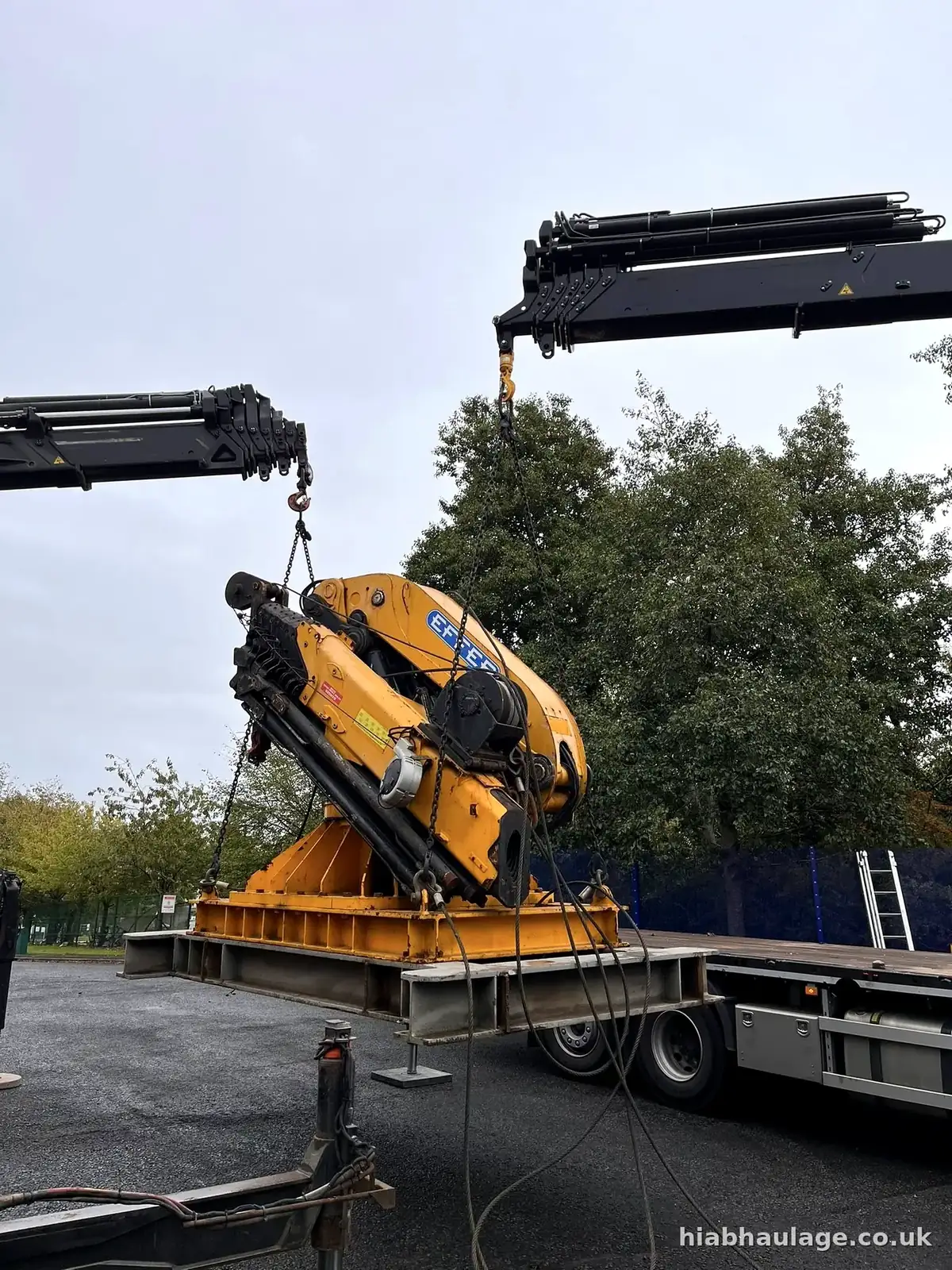Two HIAB booms lifting a yellow machine base off a trailer