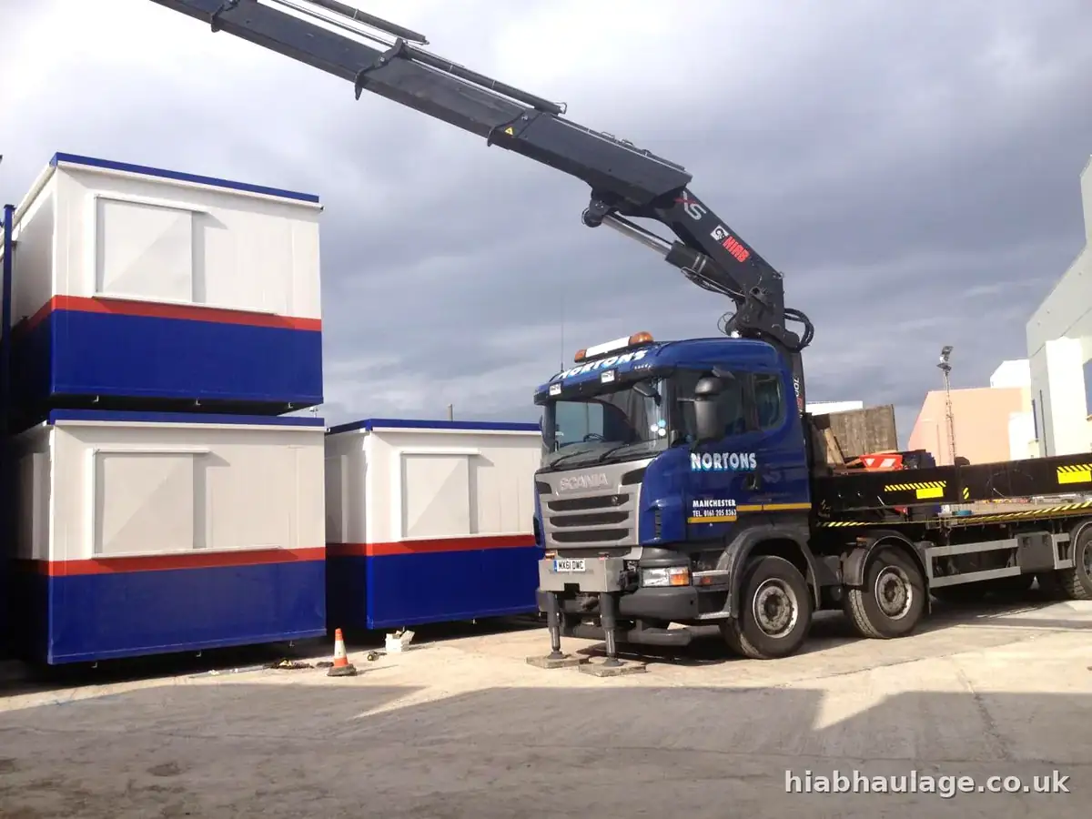 Scania rigid HIAB with its boom raised beside stacked blue and white cabins in a yard Scania rigid HIAB with its boom raised beside stacked blue and white cabins in a yard